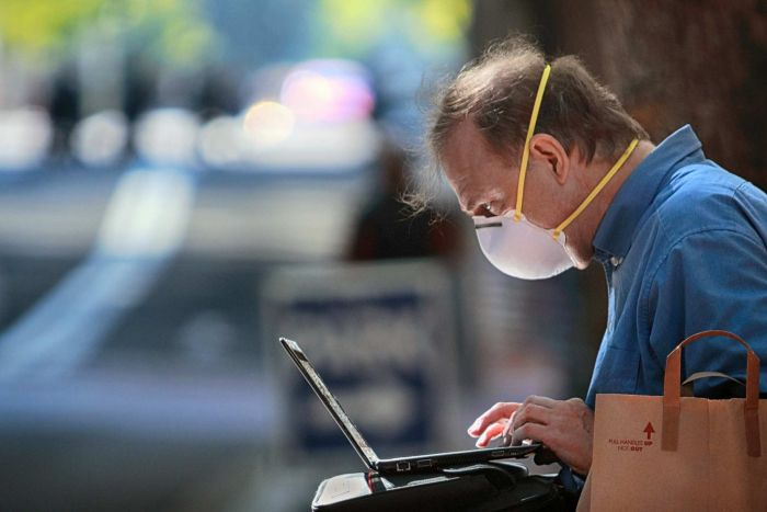 A man sitting on a bench outside wears a face mask while working on a laptop.